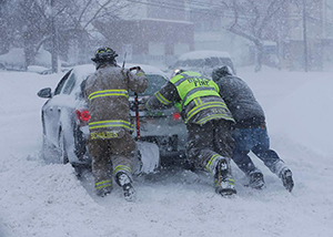 An image of three fire fighters pushing a car in a foot of snow during a snowstorm. 