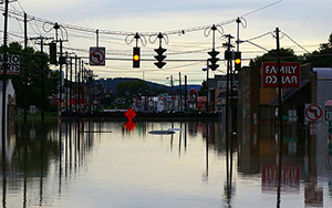 An image of a flooded street with water reaching halfway up houses and a traffic light with the yellow light lit up. 