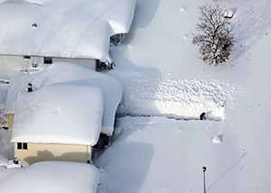 An aerial view of houses covered in snow and a person shoveling out a path when the snow is taller than them. 